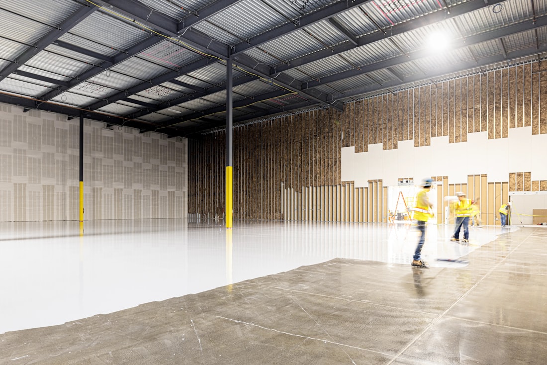 The interior of a large warehouse, with workers in high-visibility vests and a freshly cleaned, glossy floor beside bare concrete.