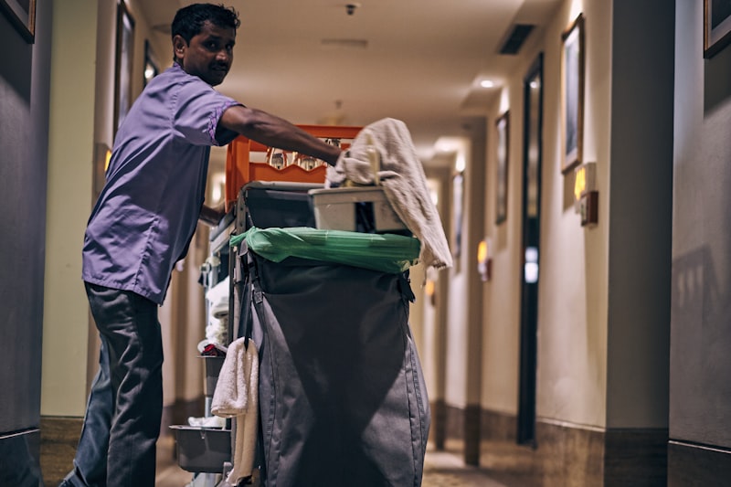 A cleaner pushing a fully stocked housekeeping trolley along a commercial corridor.