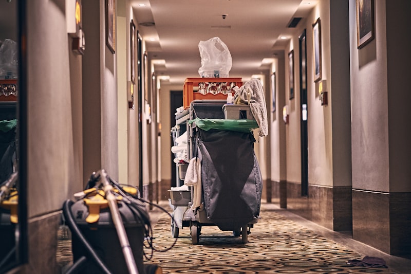 A housekeeping trolley and vacuum on patterned carpet in a long hotel corridor.