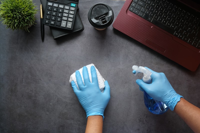Gloved hands wiping down an office desk beside a laptop with cleaning spray.