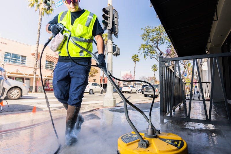 A worker in hi-vis using a high-pressure surface cleaner on a commercial footpath.
