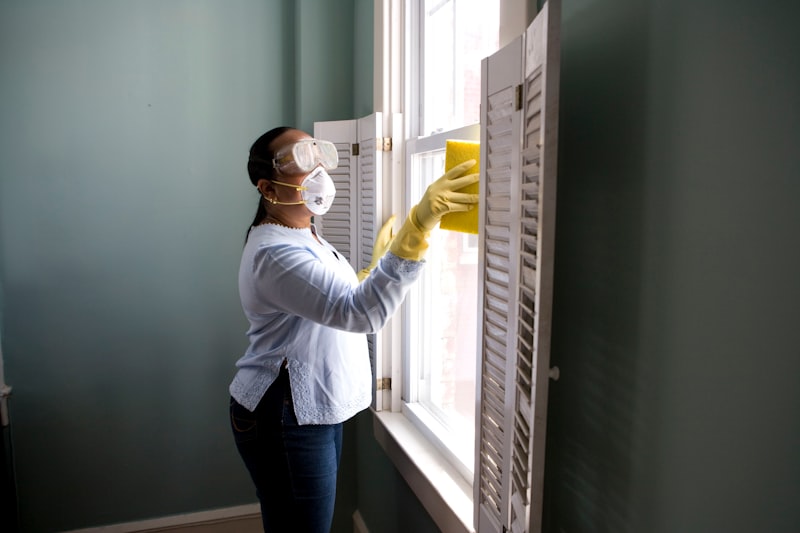A cleaner in gloves and a mask wiping down window shutters.