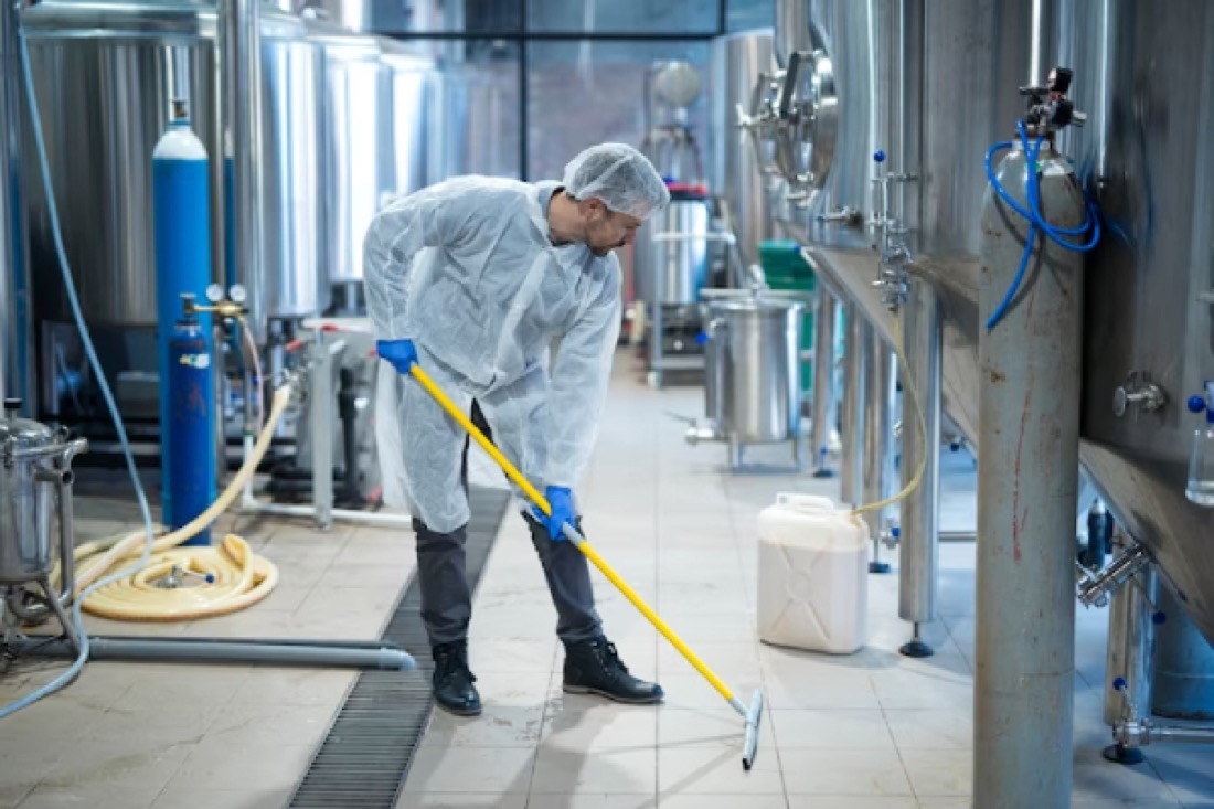 A cleaner in white protective coveralls and hairnet mopping the floor of an industrial food production facility.