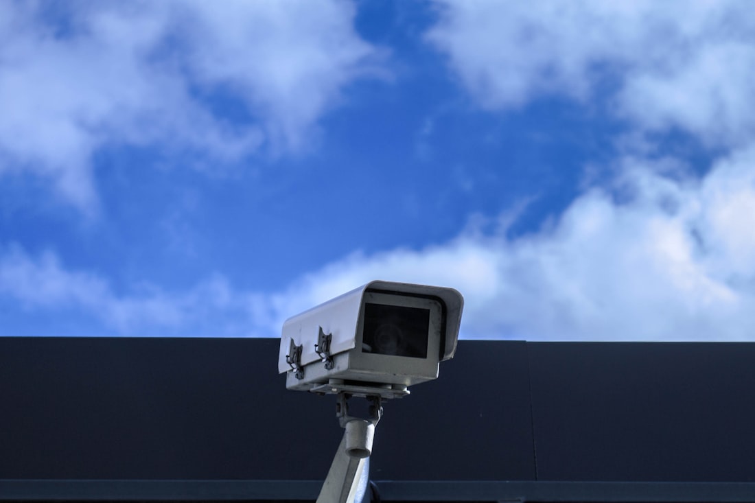 A CCTV surveillance camera mounted on the corner of a building against a blue sky.